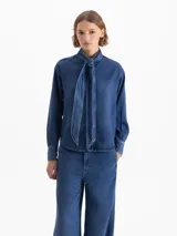 Camisa azul de manga larga confeccionada en 100% Tencel, con lazo en el cuello y acceso mediante broches dorados en la cartera delantera.