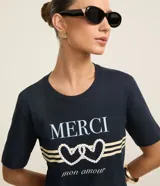 Remera azul marino de algodón con cuello redondo y mangas cortas. Estampa frontal con la palabra "Merci" en color crema, dos corazones entrelazados con detalles de perlas y la frase "mon amour" en cursiva.