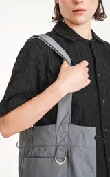 Bolso tipo tote bag color gris, confeccionado en microfibra técnica de secado rápido. Cuenta con un compartimento principal con elástico ajustable, un bolsillo interno con cierre y un bolsillo externo con velcro.