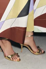 Sandalias doradas de tacón fino con tira delantera con detalle de lazo y tira con hebilla al tobillo.