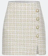 Minifalda de tweed con estampado a cuadros en tonos blanco y dorado, con tres botones decorativos dorados y blancos en el lateral y abertura frontal.