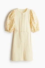 Vestido color crema a la rodilla, de tejido liviano de algodón con cuello redondo, botones ocultos adelante y cinturón fino extraíble. Mangas al codo abullonadas en broderie con puños elásticos.