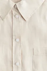 Camisa color crudo de lino liviano, con cuello clásico, botones al frente, canesú de doble capa en la espalda, hombros apenas caídos, mangas largas con puños abotonados, bolsillo superior de parche y bajo levemente redondeado.