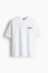 Remera blanca de algodón pesado con cuello redondo ribeteado, hombros caídos, bajo recto y estampado pequeño en el pecho. Corte oversized.