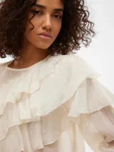 Blusa color crema con volados, cuello redondo y mangas largas con puños abotonados.