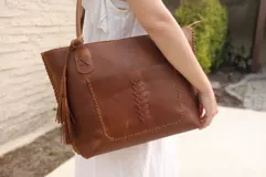 Bolso tote de cuero vacuno color marrón con bolsillo externo y detalles de costuras a la vista. Tiene doble asa de hombro con detalle de nudo y borla.