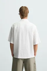 Camisa de hombre de manga corta, cuello solapa y corte relaxed fit. Tejido blanco con textura sutilmente arrugada o plisada. Cierre frontal de botonadura y bajo con aberturas laterales.