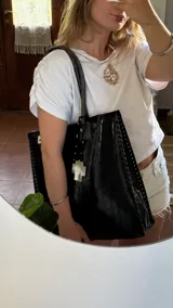 Bolso tote negro de cuero sintético con textura de cocodrilo. Presenta costuras decorativas en los laterales y un dije colgante en forma de cruz plateada con detalles de perlas.