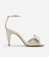 Sandalias de tacón alto color crema, con punta redonda abierta, tira fina con hebilla en el tobillo y maxi lazo decorativo en la parte delantera.