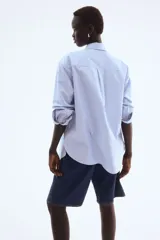 Camisa Oxford de algodón a rayas verticales celestes y blancas, con cuello clásico, botones frontales, canesú en la espalda, corte relajado, hombros caídos, mangas largas con puños sencillos y bajo redondeado con espalda más larga.