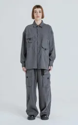 Camisa manga larga color gris grafito, estilo técnica con múltiples bolsillos delanteros y un bolsillo en la parte inferior de la espalda. Tiene botones en la cartera y las mangas, y calce oversize.