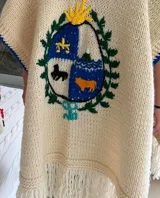 Ruana de lana merino color crema, tejida y bordada a mano, con el escudo de Uruguay en la espalda y flecos en el ruedo.