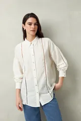 Camisa blanca de lino con botones de nácar, corte recto, cuello clásico y mangas largas con puños abotonados. Presenta detalles de calados tipo vainillas en el frente y las mangas.