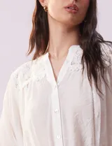 Blusa color beige de gasa con cuello a la base, escote en V y abertura central con botones. Presenta detalle de crochet en hombros y espalda. Manga 3/4 amplia con puño y botón.