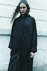 Parka larga negra con capucha ajustable con cordones, mangas largas con puños elásticos, bolsillos laterales y de plastrón en el pecho, bajo ajustable con cordón elástico y cierre frontal con cremallera metálica.