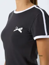 Camiseta negra de corte cropped con cuello redondo y mangas cortas con ribete blanco. Estampado pequeño de moño blanco en el pecho.