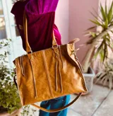 Bolso de mano grande de cuero color marrón, con doble asa corta y correa larga desmontable. Tiene cierre superior y detalles de flecos en el frente.