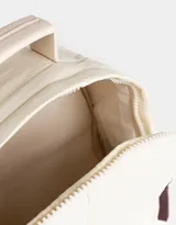 Mochila color crema de tela acolchada con pespuntes verticales. Tiene un bolsillo frontal con cierre, doble cierre superior, manija superior, correas regulables y bolsillos laterales. Incluye un charm colgante de corazón.
