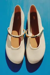 Zapato tipo bailarina color crema con acabado en punta redonda y tira sobre el empeine con aplique metálico dorado.