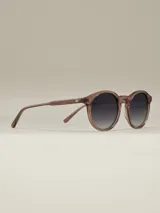 Lentes de sol de montura redonda en tono marrón rosado translúcido, con lentes oscuros degradados de negro a gris claro.