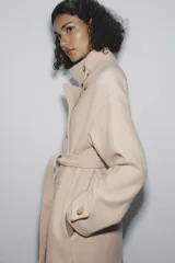 Abrigo largo color beige confeccionado en hilatura con lana. Tiene cuello subido, manga larga con trabillas, bolsillos laterales y cinturón ajustable con lazada. El cierre frontal es cruzado con botones.