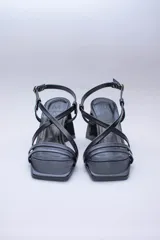 Sandalias de tacón alto color violeta metalizado, con tiras finas cruzadas en el empeine, puntera cuadrada y cierre con hebilla en el tobillo. El tacón es geométrico con forma de triángulo invertido.