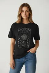 Remera de algodón negra de manga corta y corte holgado, con cuello a la base. Presenta un estampado frontal en color claro con motivos celestiales, incluyendo un sol central, manos y texto.