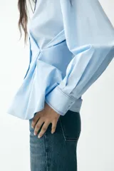 Camisa celeste de corte holgado, con cuello solapa, manga larga y cierre frontal con botones. Presenta detalle de cintura ajustable con botones.