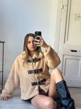 Campera corta color beige de tela tipo peluche con cuello solapa y cierre frontal con presillas de cuero sintético y hebillas metálicas.