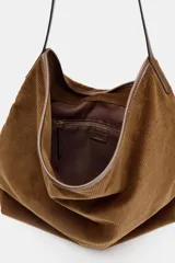 Bolso tipo shopper de gran tamaño, confeccionado en pana marrón con textura acanalada. Presenta un asa de hombro de cuero marrón y un detalle de tira ajustable con hebilla metálica en la parte frontal.
