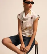 Blusa sin mangas de tela satinada color blanco, con cuello drapeado, lazo lateral y estampado de manchas en tonos rosados, amarillos y verdes.