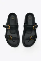 Sandalias planas negras con dos tiras ajustables con hebillas doradas y punta redonda.