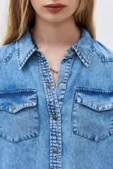 Camisa de denim celeste con cuello clásico, cierre frontal con botones a presión, dos bolsillos con solapa en el pecho y mangas largas con puños abotonados.