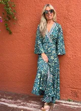 Vestido largo estilo cruzado (wrap dress) de manga larga acampanada, con estampado floral en tonos verde petróleo, amarillo pálido y celeste. Presenta un ribete decorativo en el escote y el borde del cruce.