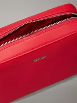 Cartera cruzada tipo 'camera bag' de Calvin Klein, color rojo vibrante, con forma rectangular y esquinas redondeadas. Presenta un acabado liso y el logo metálico de la marca en el centro frontal. Incluye herrajes metálicos plateados y una correa ajustable con detalles en tela.