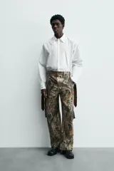 Camisa blanca de algodón popelín, corte relajado, con cuello solapa y pechera frontal plisada. Las mangas terminan en puños ajustables con lazos largos estampados en color marrón con patrón circular.