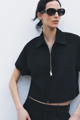 Chaqueta corta negra de cuello solapa y manga corta. Cierre frontal con cremallera metálica y bolsillos de ojal. Zara Woman Collection.
