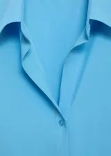 Camisa celeste de tejido fluido con cuello camisero, cierre frontal con botones y mangas largas con puños abotonados.