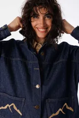 Camisa corta de jean azul con terminación redondeada en el ruedo. Presenta dos bolsillos plaqué bordados con motivo de montaña y detalle de pespunte a contratono.