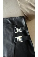 Minifalda negra de cuero sintético con corte irregular y detalle de hebillas decorativas en el lateral.