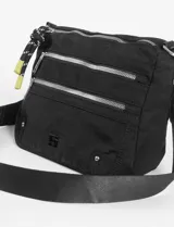 Bandolera negra de tela con múltiples cierres horizontales plateados. Incluye una correa ajustable y detalles colgantes en los tiradores de los cierres, uno de ellos es una borla trenzada blanca y negra y otro es un tirador pequeño de color amarillo.