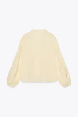 Blusa color crema de escote pico con volantes y manga larga. Detalle de jaretas y entredoses a tono. Cierre frontal con botones forrados a tono.