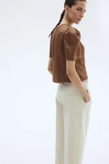 Jean tobillero de denim de algodón rígido color beige claro, con corte estándar de cadera a muslo y piernas con volumen de diseño curvo que se ciñen en el bajo. Presenta tiro alto, cierre con botón, bolsillos al bies, bolsillos decorativos traseros y pliegues delanteros y traseros.