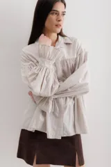 Camisa gris a rayas verticales blancas, de corte amplio, cuello alto con lazo y mangas largas abullonadas con puños elásticos.