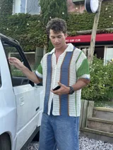 Camisa de manga corta tejida a crochet, con cuello polo y botones frontales. Presenta un diseño de rayas verticales en blanco, azul, verde y marrón.