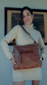 Cartera marrón de símil cuero con bolsillo frontal con tapa y cierre. Tiene apertura superior con cierre, división interna, bolsillos internos y bolsillo trasero con cierre. Incluye correa larga regulable y asa de mano.