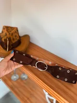 Cinturón ancho de doble capa, con capa superior de gamuza negra o marrón y capa inferior de cuero color camel. Presenta múltiples ojales metálicos y hebilla ovalada metálica de color dorado.