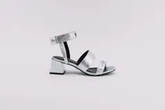 Sandalias plateadas con dos tiras horizontales sobre el pie y una tira vertical que se une a una pulsera con hebilla en el tobillo. La pulsera tiene ojales y tachas combinadas. El taco es cuadrado y de 5 cm.