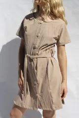 Vestido corto color beige, de tusor 100% algodón, con botones frontales, cuello redondo y mangas cortas. Incluye cinturón de la misma tela.