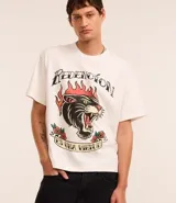 Remera color crema de corte recto, con cuello redondo y mangas cortas. Presenta un estampado estilo tatuaje con un felino en llamas y la frase "Redención es una virtud".